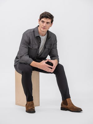 Model wearing Black Pace Tapered Chino and Charcoal Tweed Fusion Overshirt and Pale Grey Heather Composite Merino Tee sitting on a block with sleeves rolled and hands clasped
