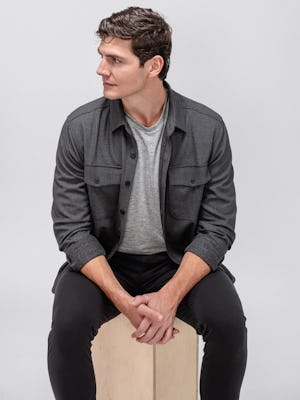 Model wearing Black Pace Tapered Chino and Charcoal Tweed Fusion Overshirt and Pale Grey Heather Composite Merino Tee sitting on a block with sleeves rolled and hands clasped