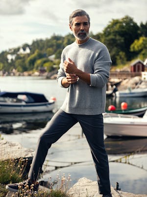 model wearing mens atlas waffle roll neck sweater grey heatherfull body hands touching one leg on small rock boats background