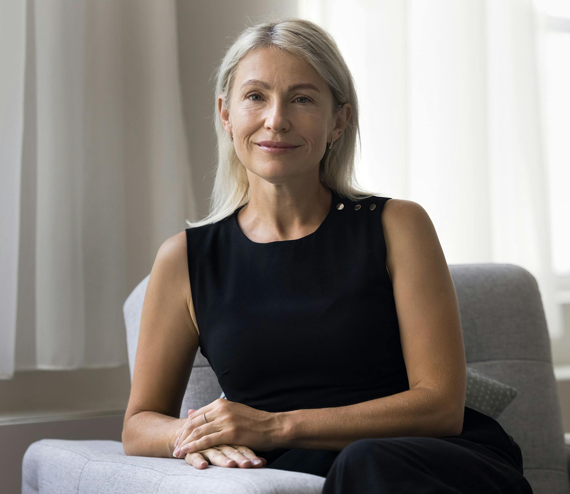 woman in black tank top and black pants leans on grey couch arm