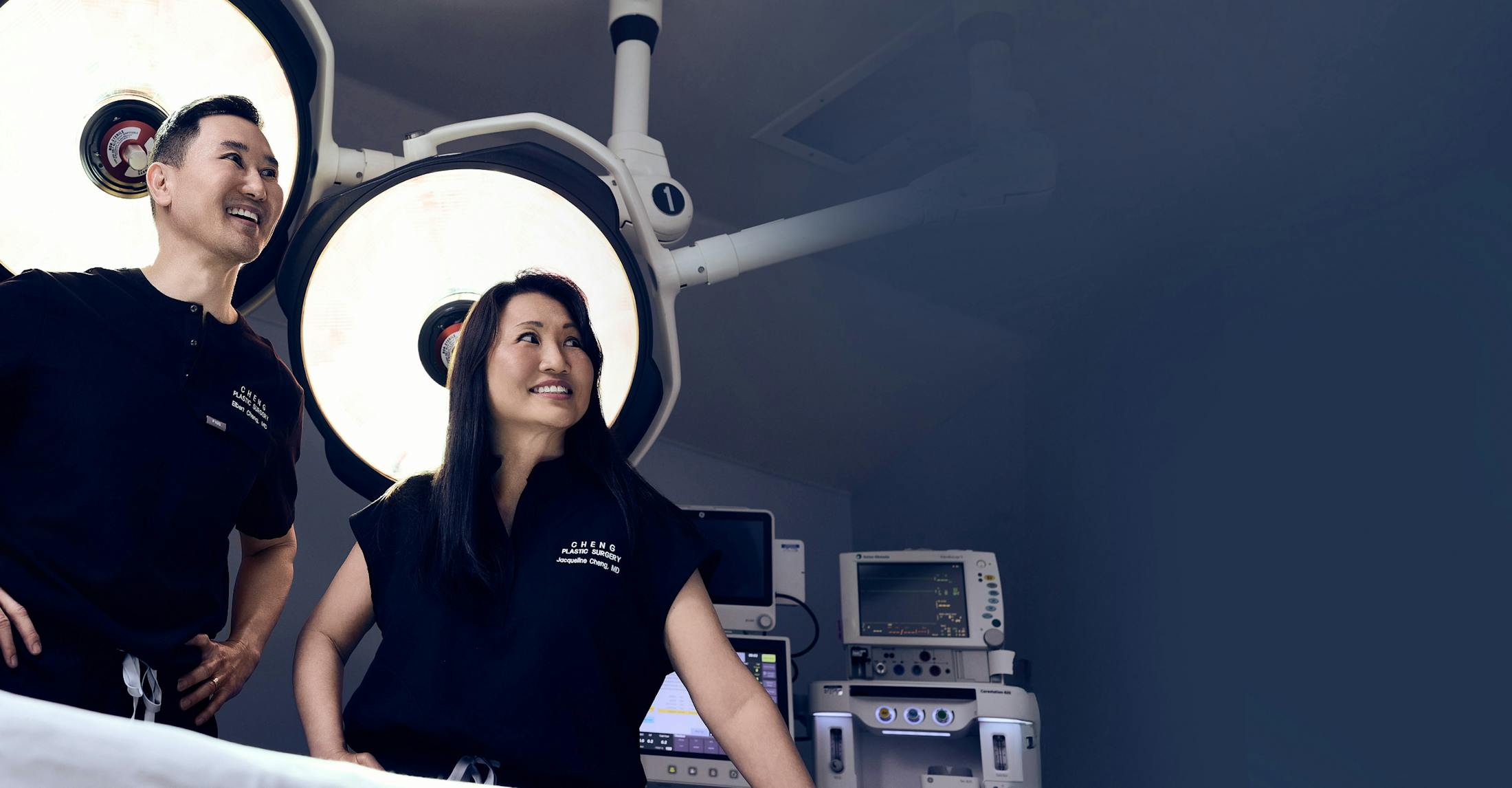 Dr. Elbert Cheng and Dr. Jacqueline Cheng in scrubs standing in front of surgical lights