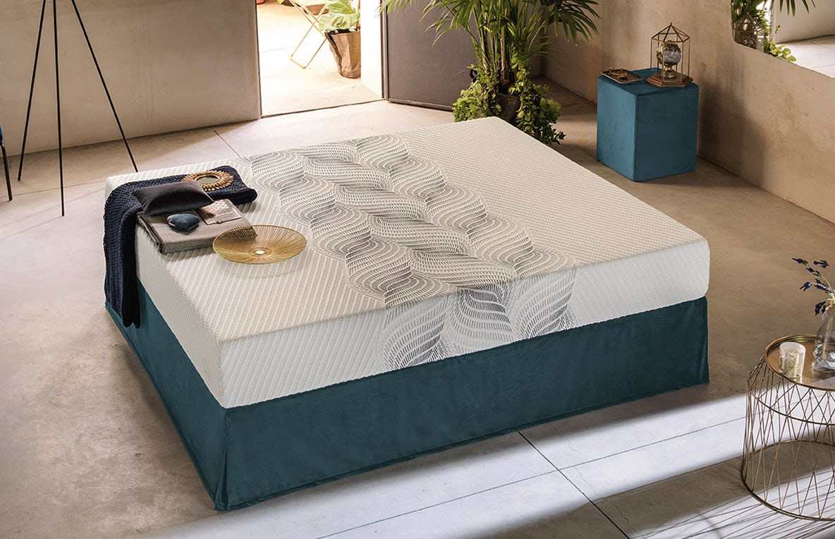 Materasso bianco con decorazione rigata su base blu scuro, coperta e cuscino grigi appoggiati sopra in ambiente moderno con…