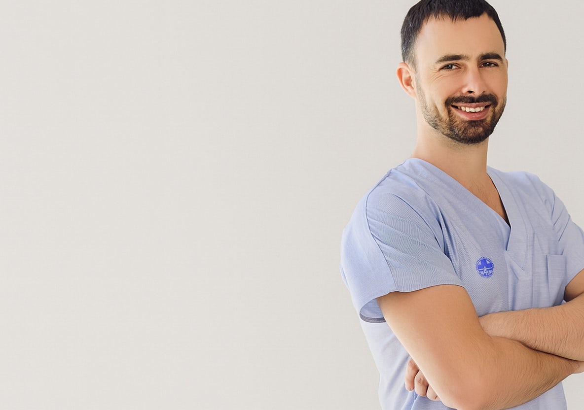Medico uomo sorridente con camice azzurro e braccia incrociate su sfondo chiaro neutro.