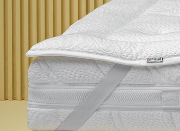 Materasso bianco con coprimaterasso imbottito fissato da elastico su struttura letto in legno chiaro, sfondo a listelli…