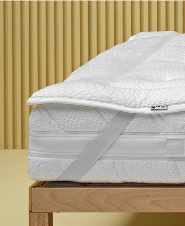 Materasso bianco con coprimaterasso imbottito fissato da elastico su struttura letto in legno chiaro, sfondo a listelli…