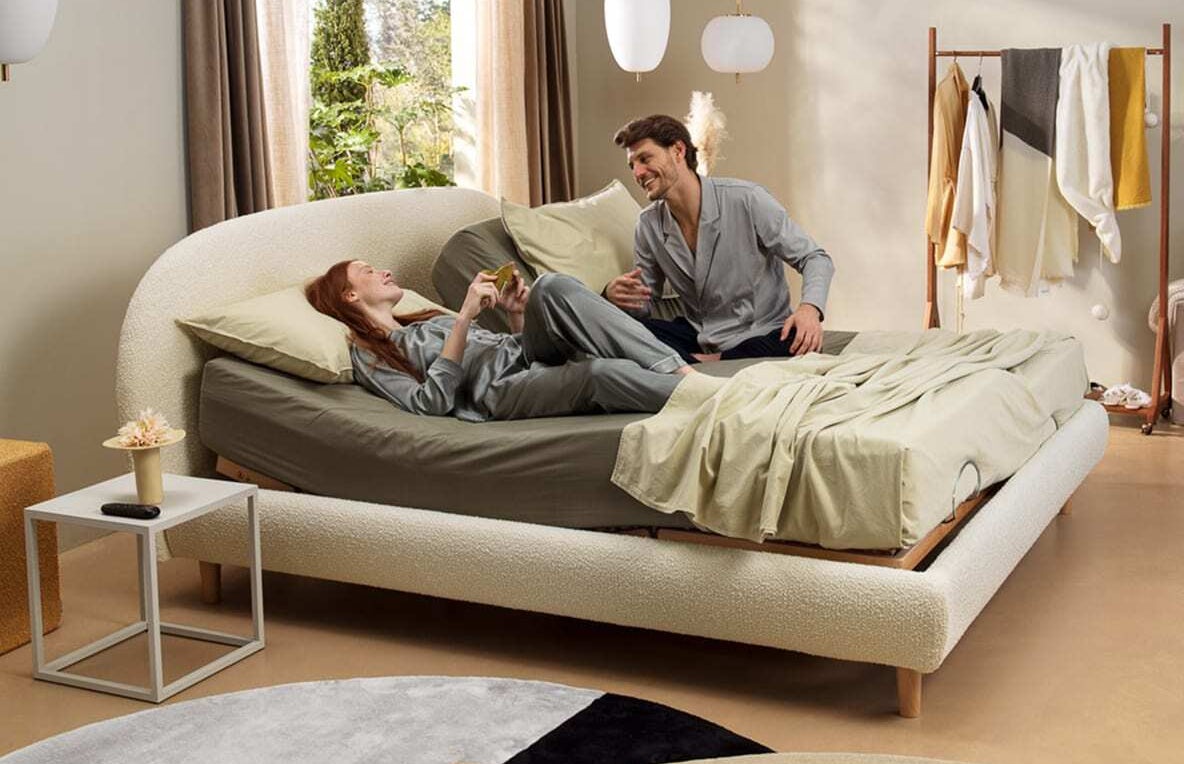 Coppia in pigiama grigio chiaro su letto moderno con testiera curva, accanto finestra e portabiti con vestiti colorati.