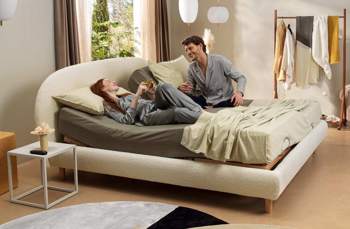 Coppia in pigiama grigio chiaro su letto moderno con testiera curva, accanto finestra e portabiti con vestiti colorati.