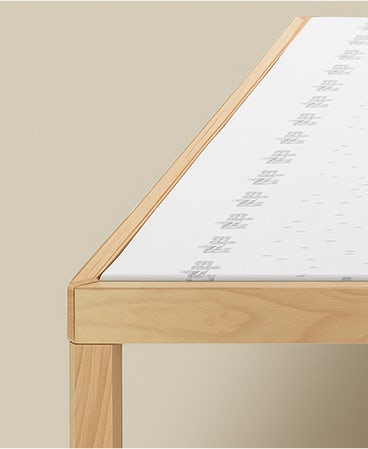 Particolare angolo di un letto con struttura in legno chiaro e materasso bianco con scritta grigia ripetuta.