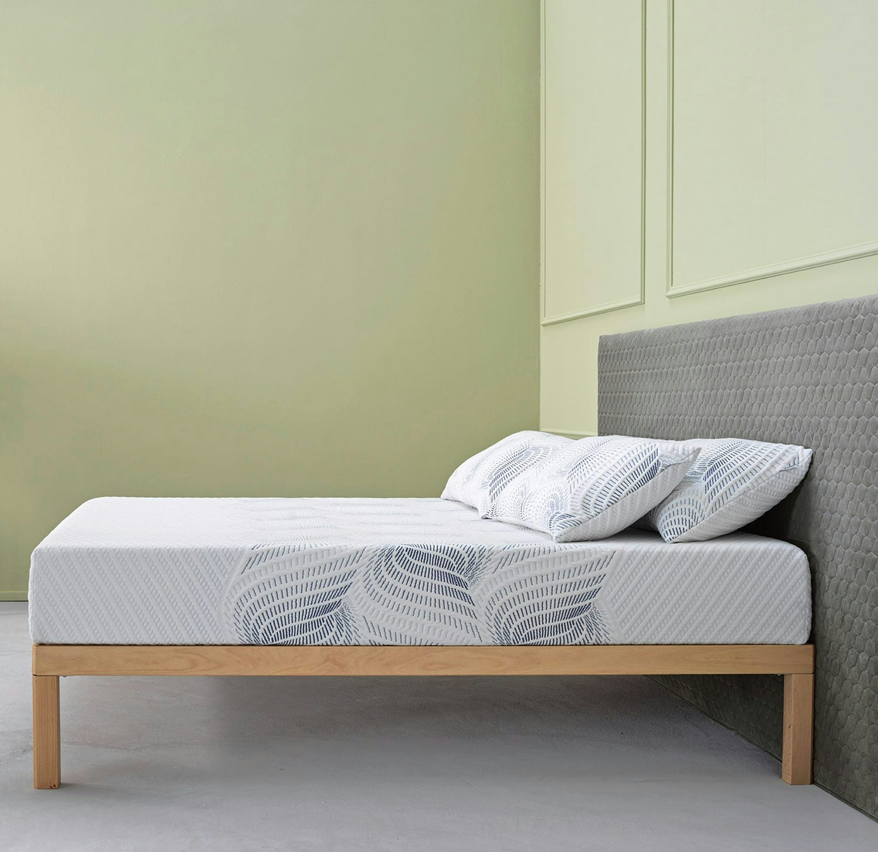 Letto con base in legno chiaro, materasso bianco con motivo blu e testiera imbottita grigia in ambiente con pareti verde…