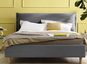Letto imbottito grigio con cuscini bianchi e giallo, parete gialla e pianta verde sullo sfondo, tappeto giallo davanti.