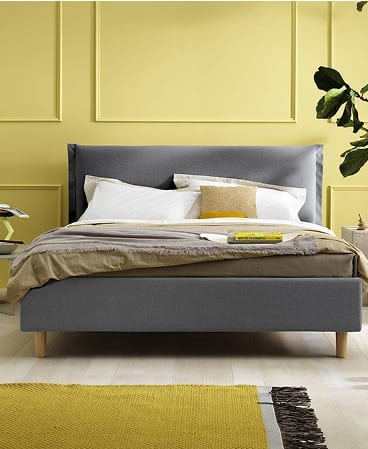 Letto imbottito grigio con cuscini bianchi e giallo, parete gialla e pianta verde sullo sfondo, tappeto giallo davanti.