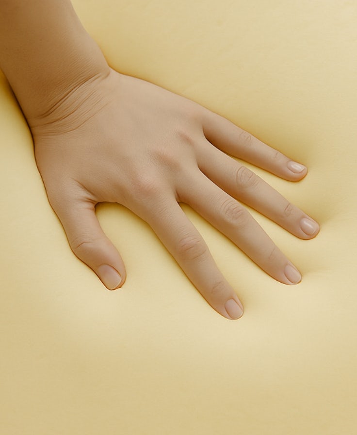 Mano femminile che preme delicatamente un materasso o cuscino memory foam di colore beige chiaro.