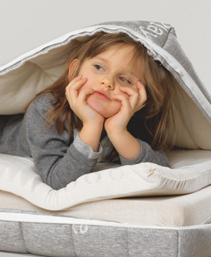 Bambina con maglione grigio sdraiata sotto un coprimaterasso grigio chiaro, appoggiando il viso sulle mani.