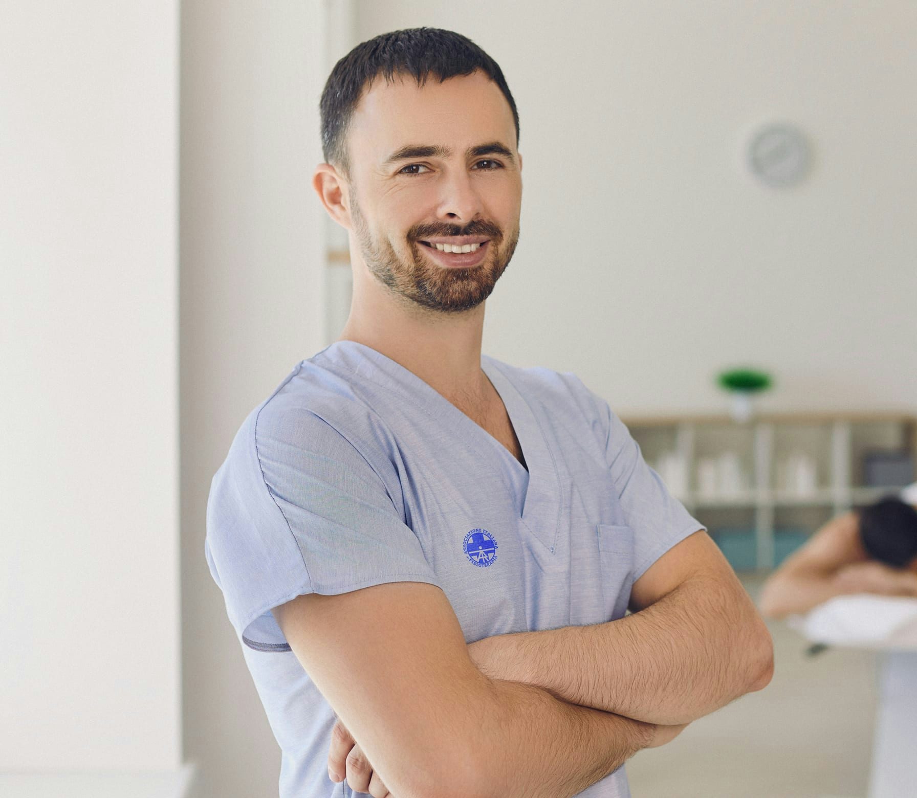 Fisioterapista sorridente con divisa blu chiaro e braccia incrociate in studio con paziente sul lettino.