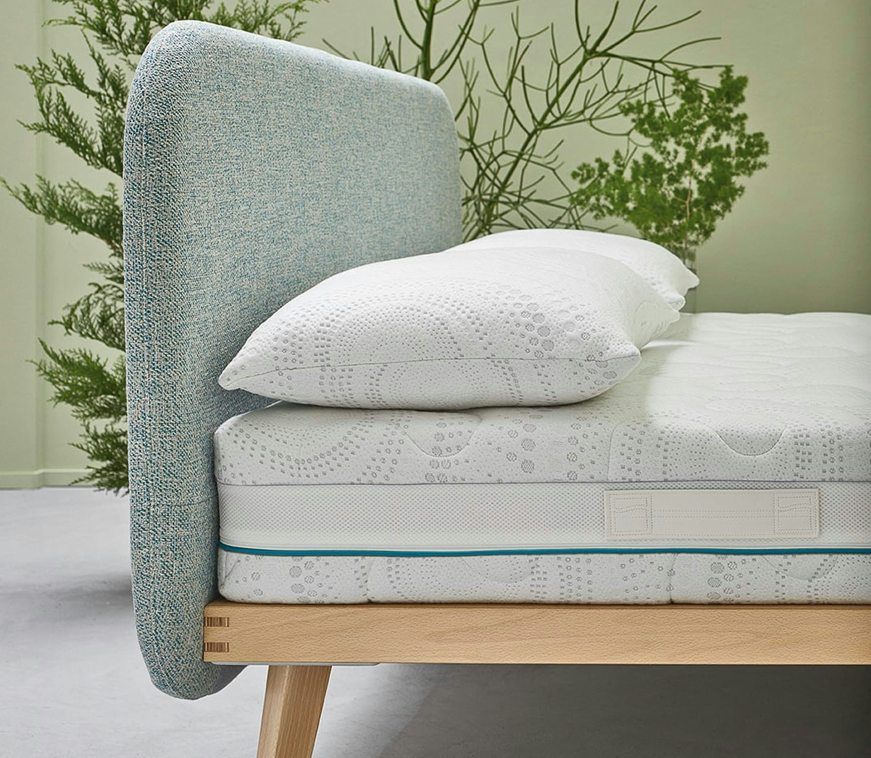 Letto con testiera imbottita azzurra, materasso e cuscino bianchi su struttura di legno chiaro con gambe inclinate.