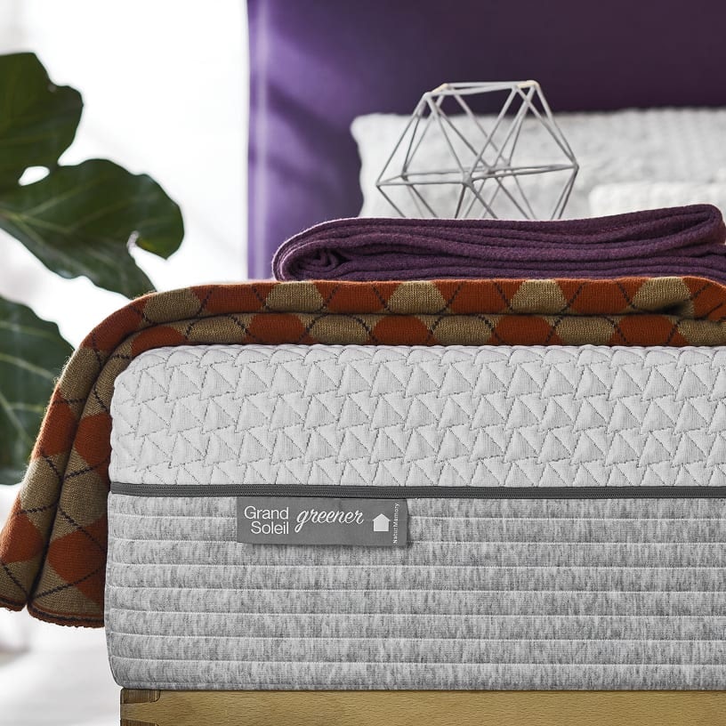 Letto con materasso Grand Soleil Greener grigio chiaro con coperta a rombi arancioni e plaid viola sopra, sfondo viola.