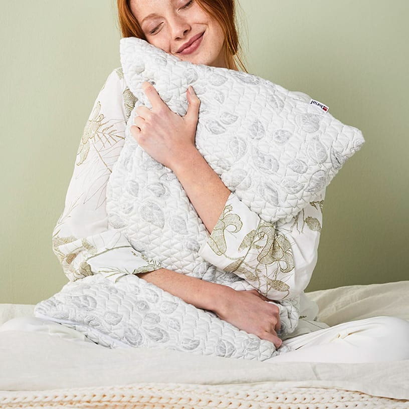 Donna con capelli rossi abbraccia due cuscini bianchi trapuntati con motivi a foglie, seduta su letto con coperta beige.