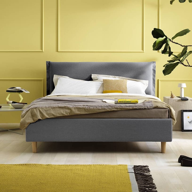 Camera da letto con letto matrimoniale grigio, pareti gialle, comodino con lampada e pianta a foglie grandi.