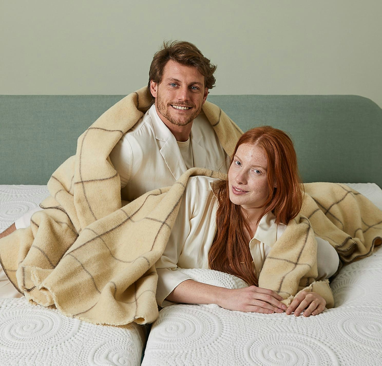 Coppia avvolta in coperta beige a quadri su un letto matrimoniale con materasso bianco e testiera verde menta
