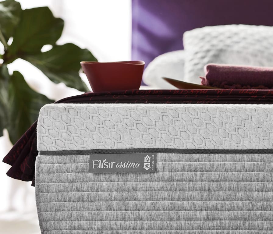 Dettaglio di materasso grigio con etichetta Elisirissimo Memory Gel, coperta viola e tazza rossa in soggiorno con pianta…