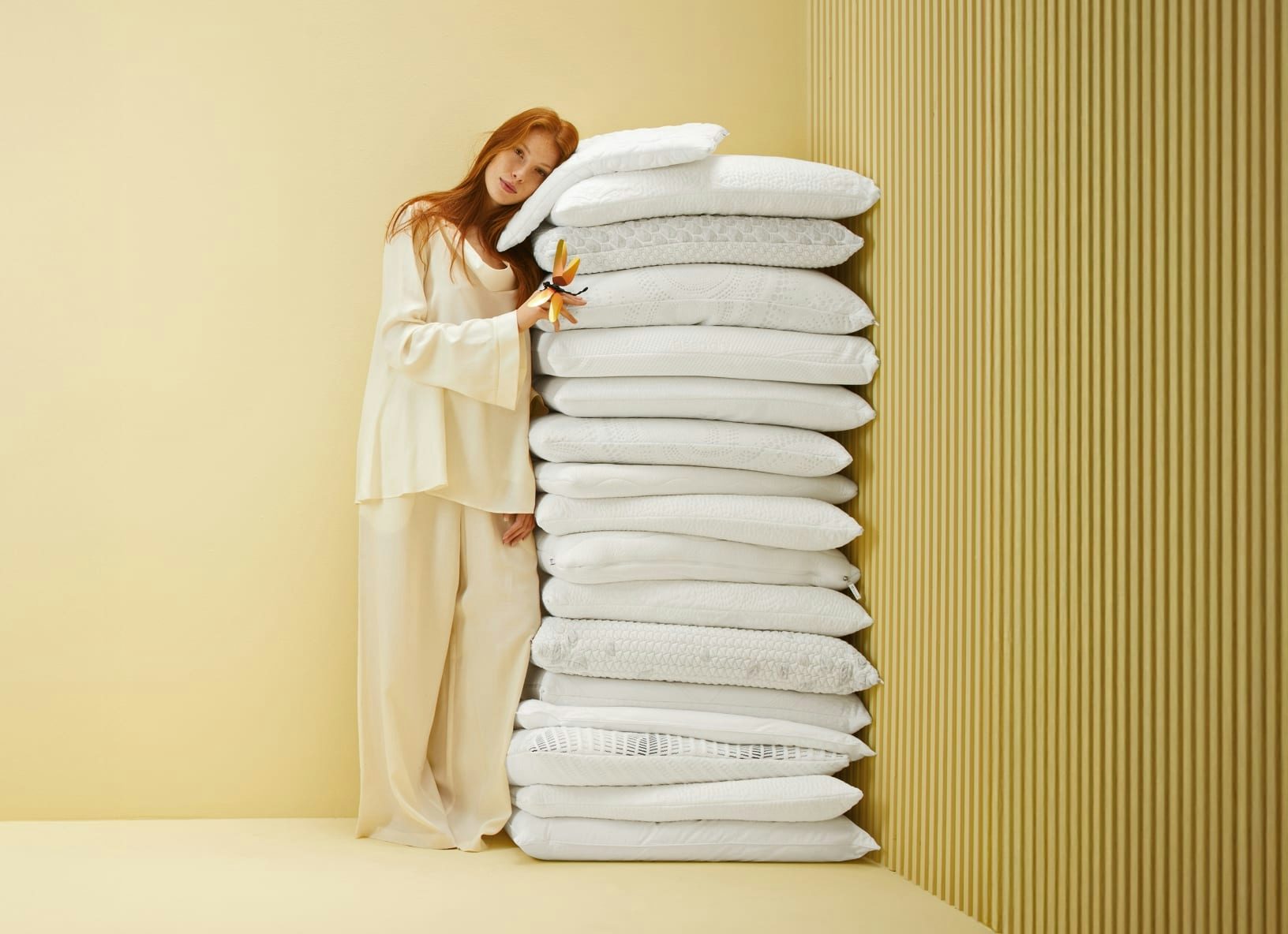 Donna con pigiamone beige appoggiata a una pila alta di cuscini bianchi contro parete beige rigata.