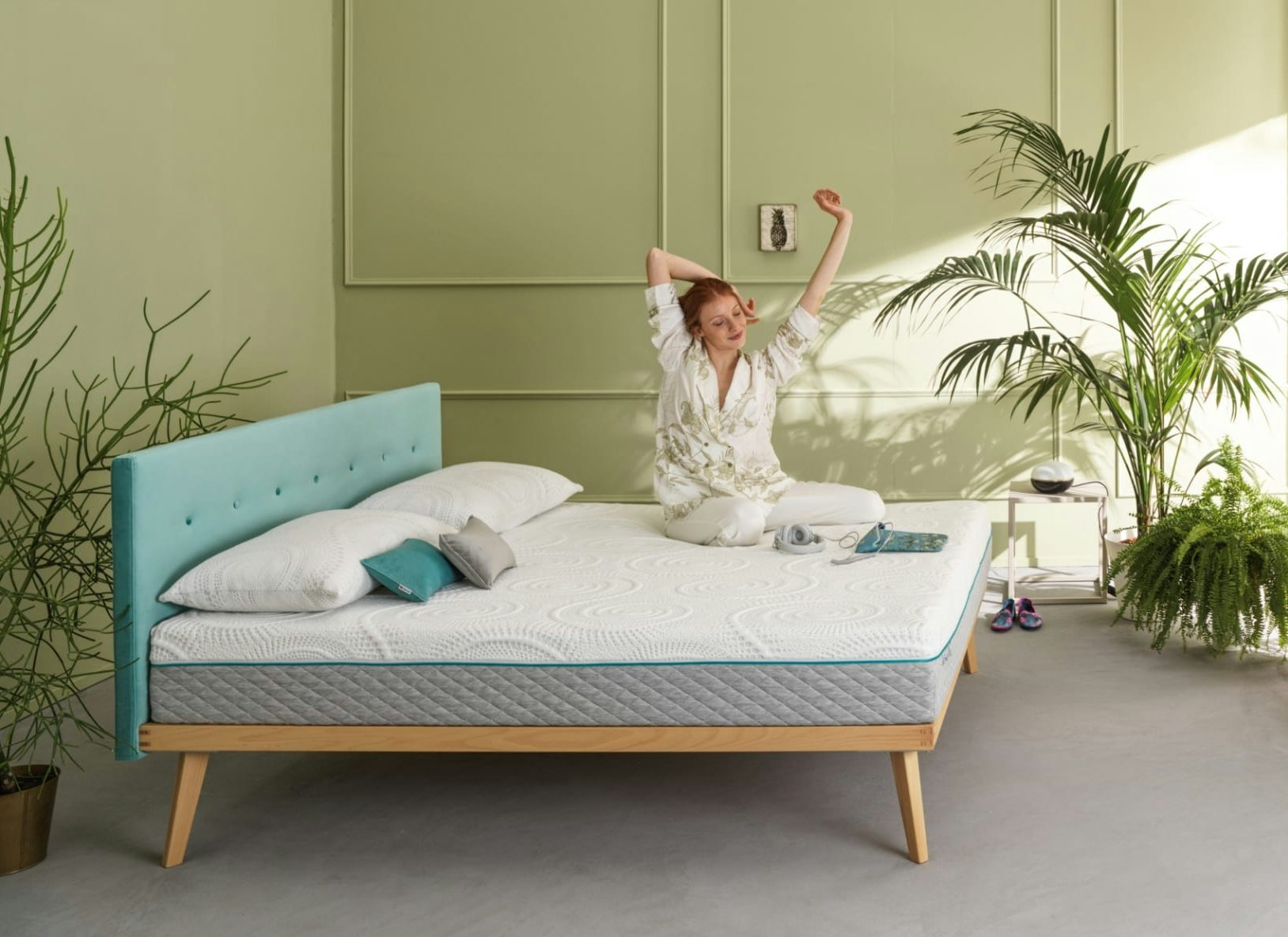 Donna rossa che si stira seduta su un letto con testiera azzurra in stanza minimalista con piante verdi e pareti salvia.