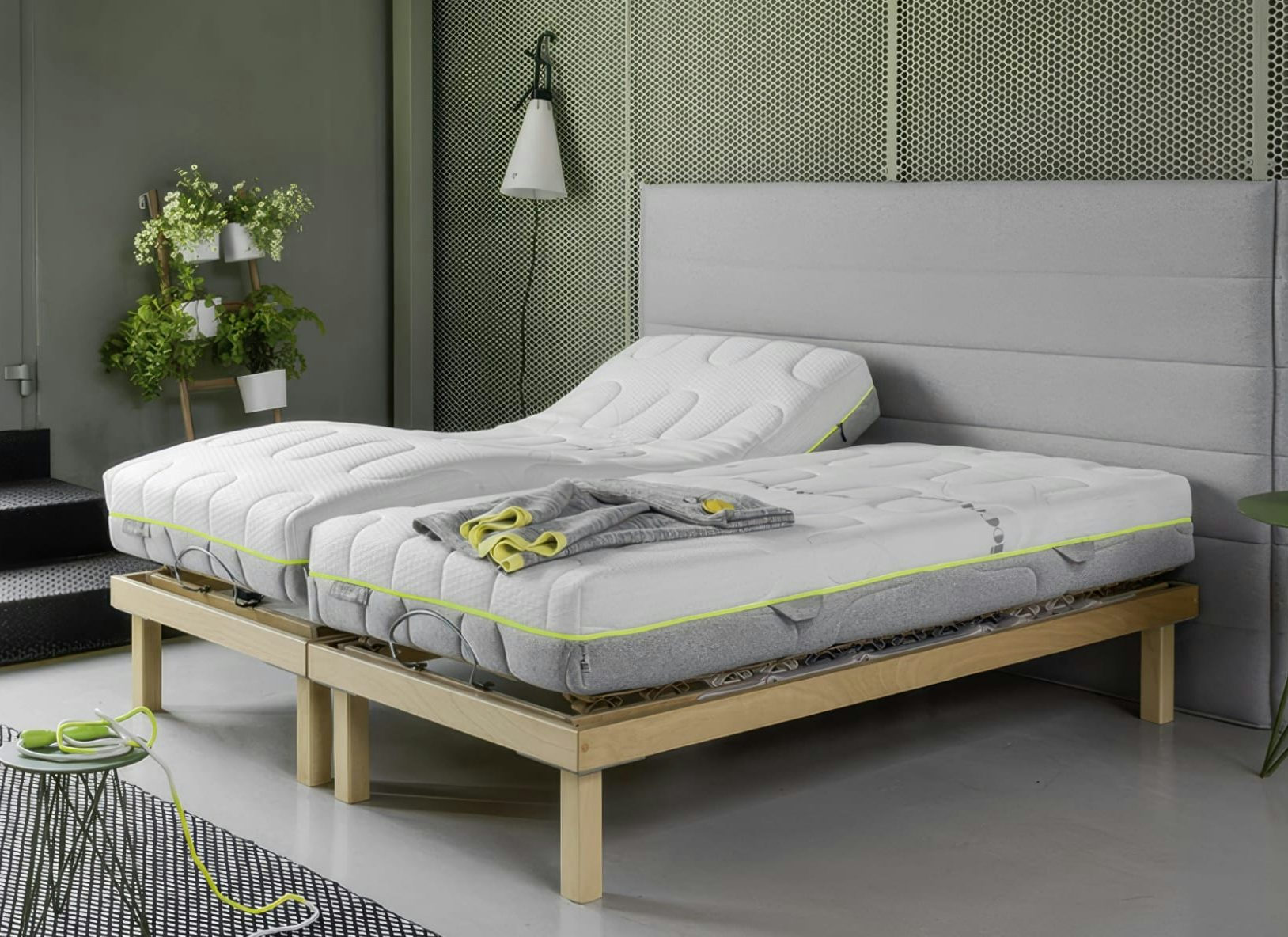 Letto matrimoniale con rete motorizzata in legno chiaro, materassi bianchi con bordo giallo fluo e plaid grigio piegato.