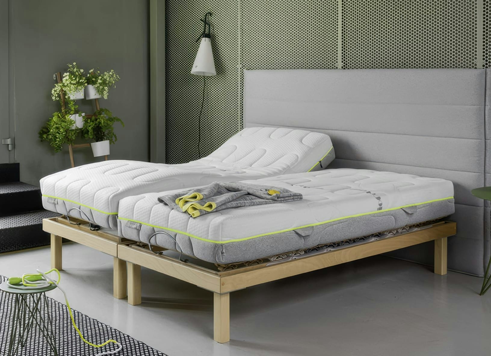 Letto matrimoniale con rete motorizzata in legno chiaro, materassi bianchi con bordo giallo fluo e plaid grigio piegato.