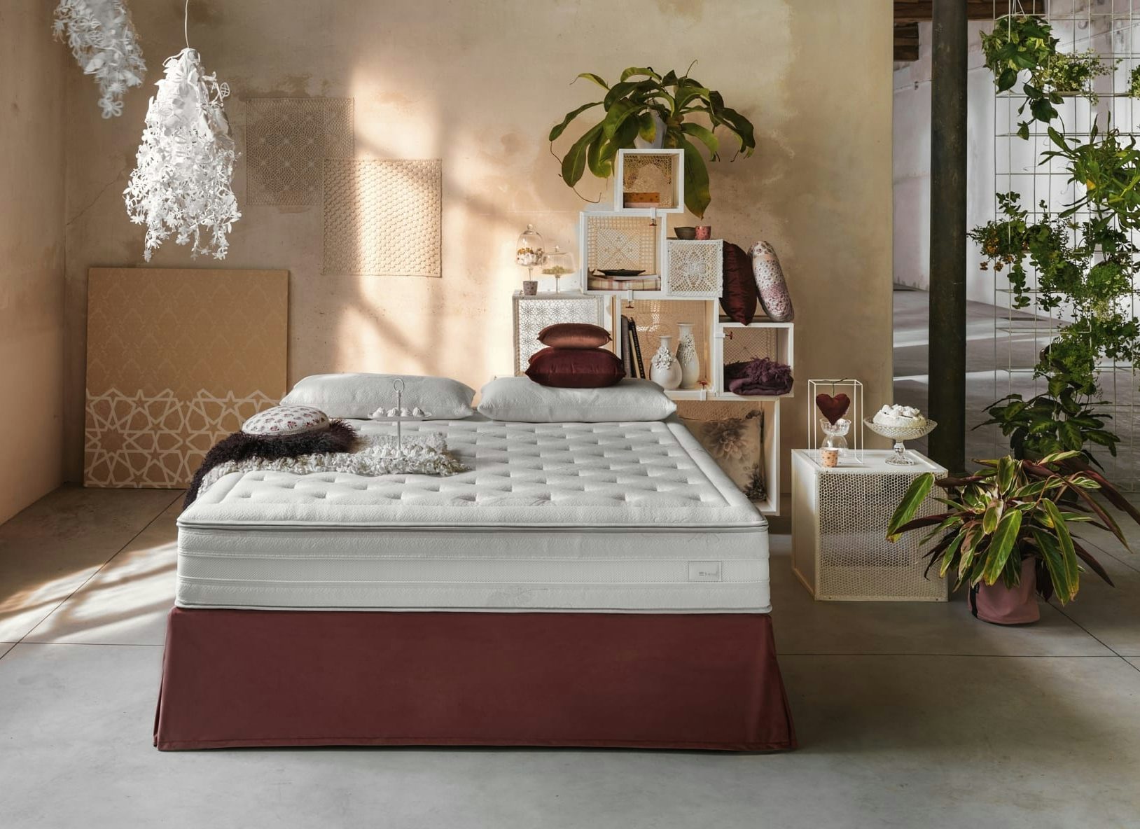 Letto matrimoniale con materasso bianco e base rossa, cuscini di velluto, scaffale cubico con piante e decorazioni, parete…