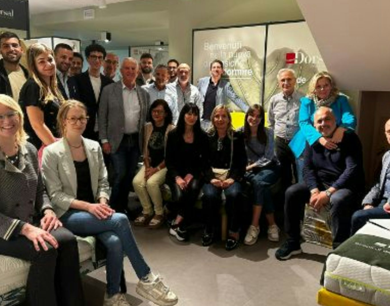 Gruppo di persone sorridenti in una stanza con materassi, sfondo con scritte benvenuti nello spazio Doze.