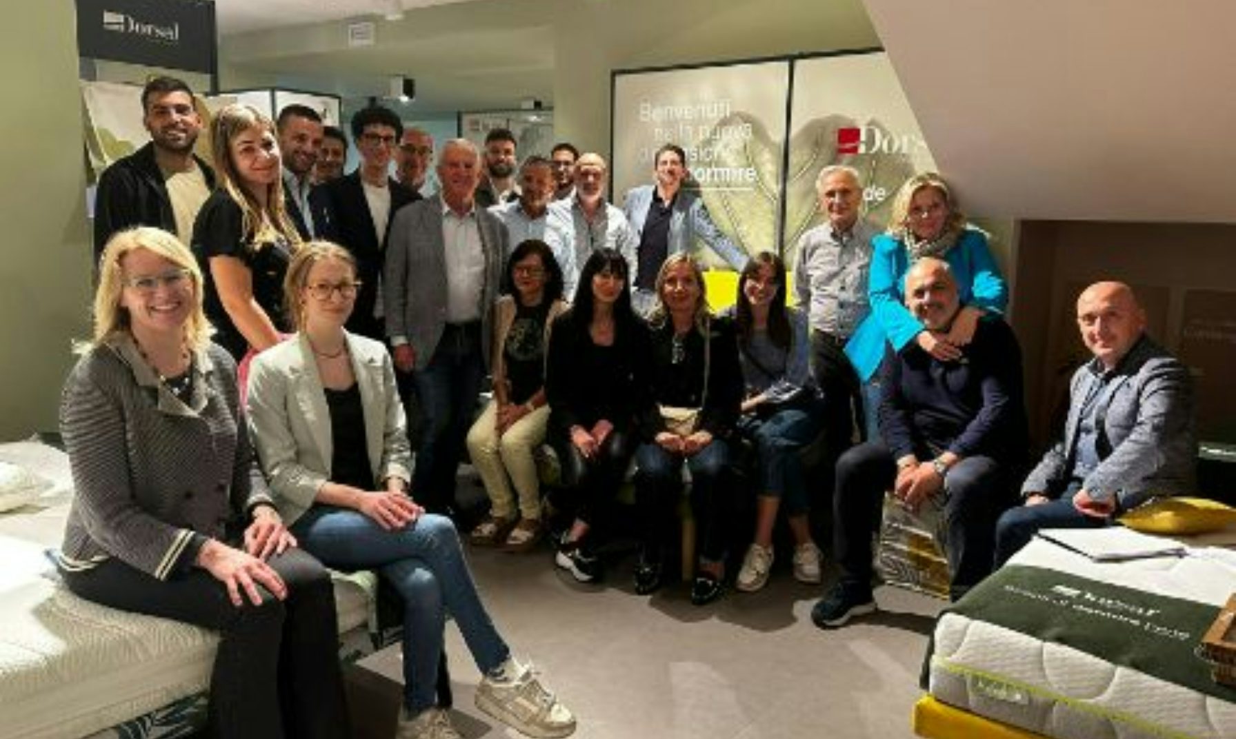 Gruppo di persone sorridenti in una stanza con materassi, sfondo con scritte benvenuti nello spazio Doze.