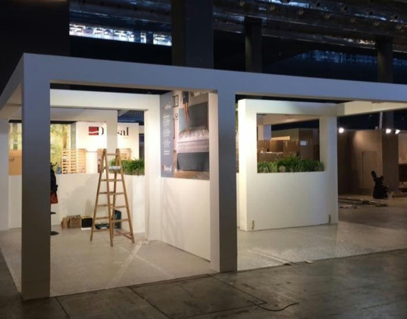 Struttura espositiva bianca in fase di allestimento con scala e piante in ambiente industriale.