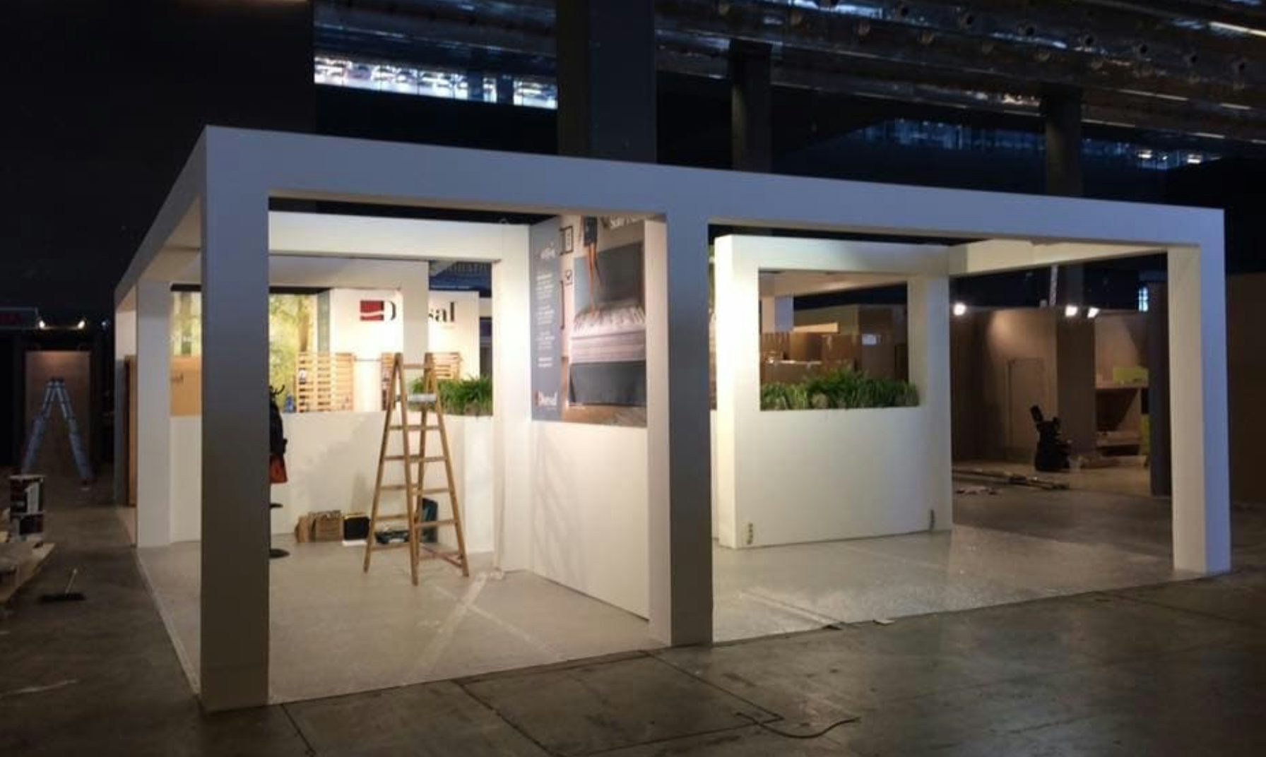 Struttura espositiva bianca in fase di allestimento con scala e piante in ambiente industriale.