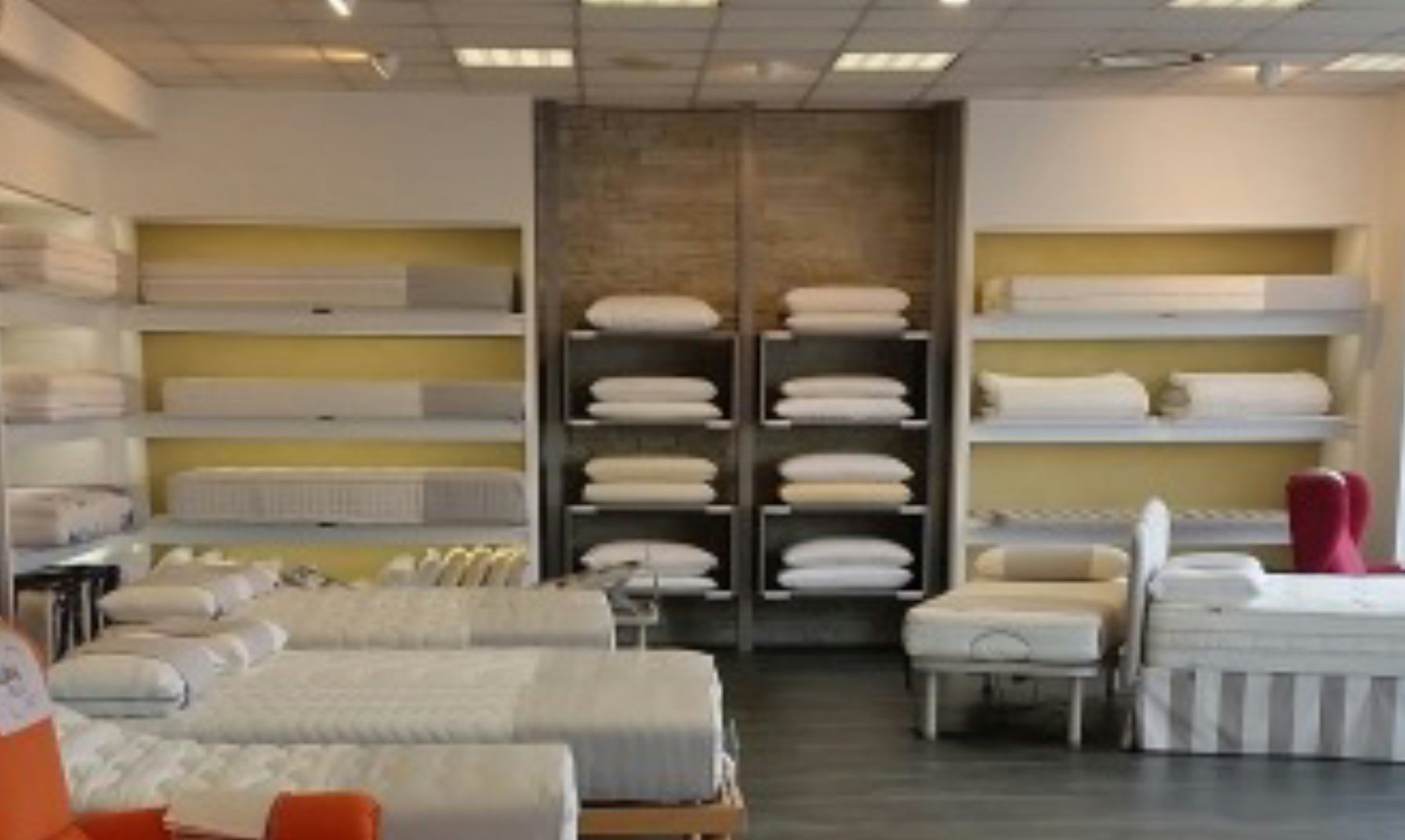 Negozio con letti matrimoniali e singoli, cuscini e reti esposti su scaffali e pavimento in legno scuro.