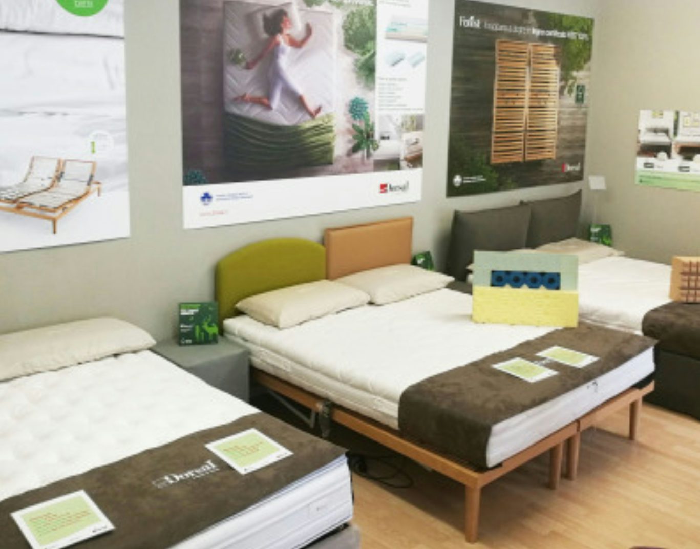 Tre letti singoli esposti in showroom con cuscini, copriletto marrone e poster informativi sulle pareti.