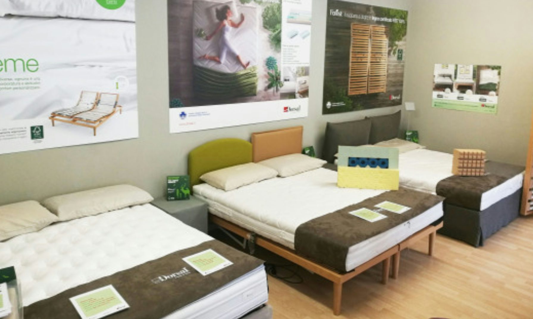 Tre letti singoli esposti in showroom con cuscini, copriletto marrone e poster informativi sulle pareti.