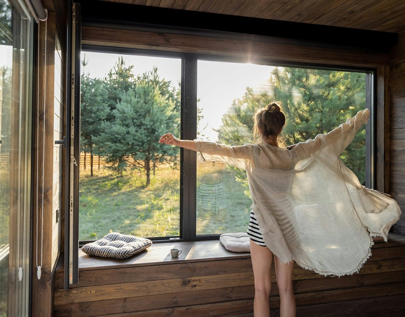 Donna con kimono leggero apre le braccia davanti a grande finestra in legno con vista su alberi al sole.