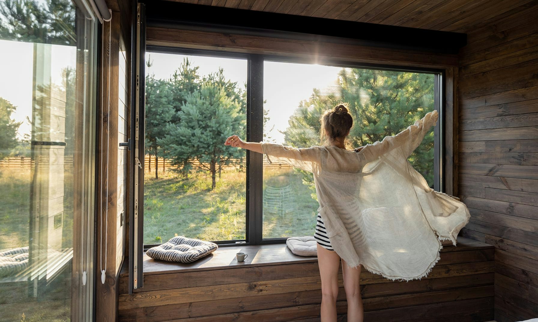 Donna con kimono leggero apre le braccia davanti a grande finestra in legno con vista su alberi al sole.