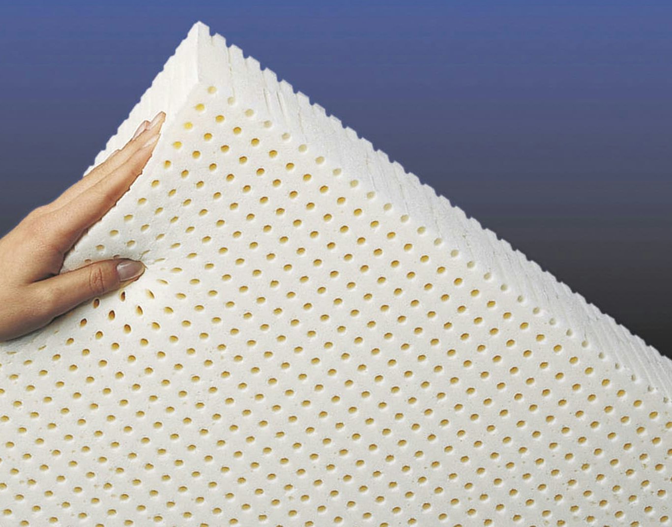 Mano che regge un materasso bianco in lattice con fori uniformi su sfondo sfumato blu e nero