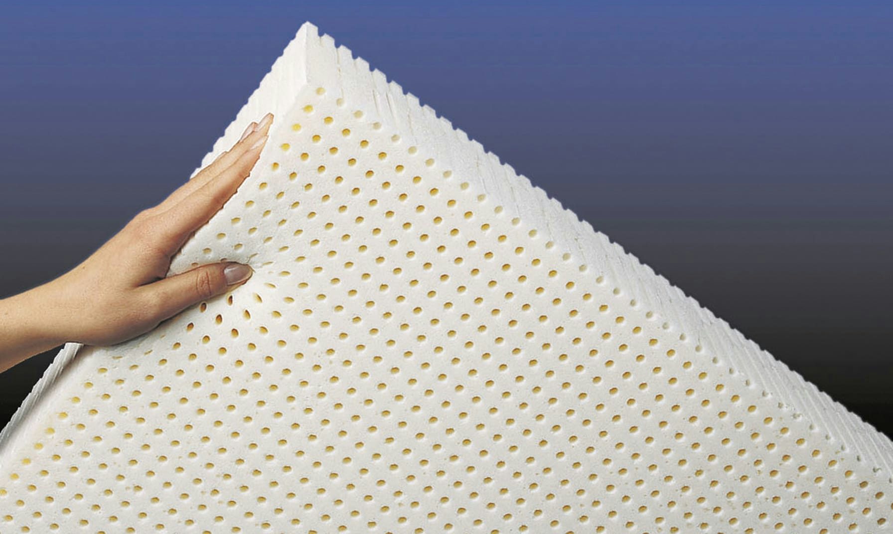 Mano che regge un materasso bianco in lattice con fori uniformi su sfondo sfumato blu e nero