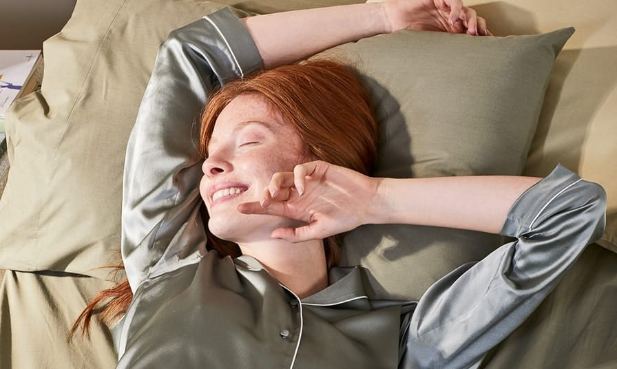 Donna con capelli rossi su cuscino verde chiaro che sorride e si stira in pigiama di raso grigio chiaro.
