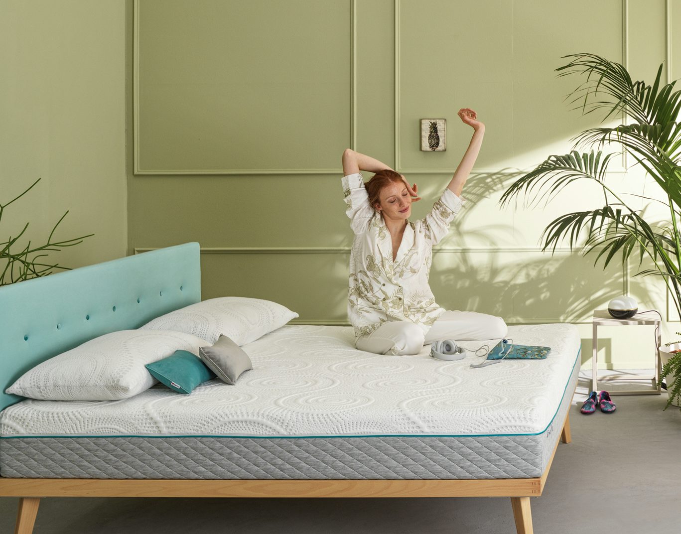 Donna con pigiama bianco che si stiracchia seduta su un letto con testiera turchese in camera verde con piante.