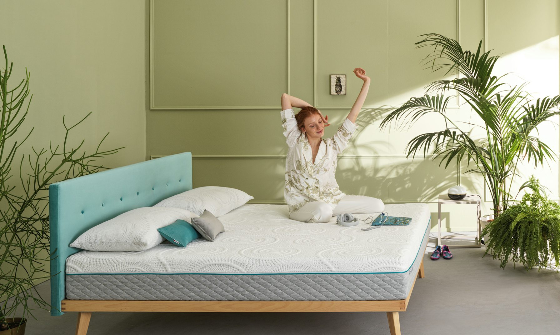 Donna con pigiama bianco che si stiracchia seduta su un letto con testiera turchese in camera verde con piante.