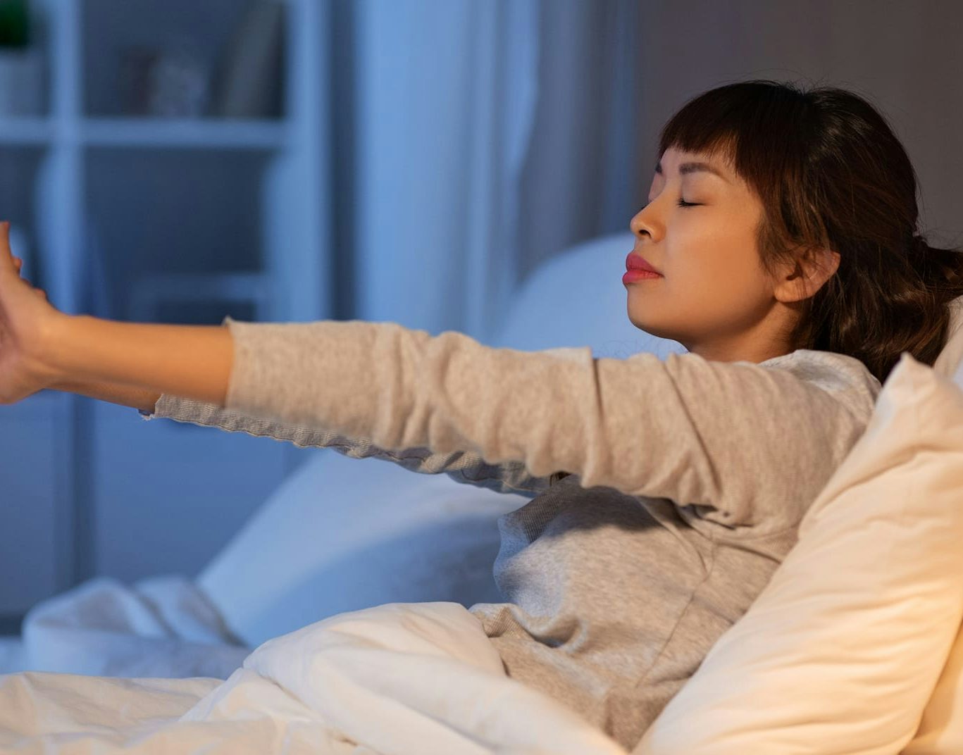 Donna asiatica che si sveglia a letto e si allunga con gli occhi chiusi, ambiente domestico notturno.