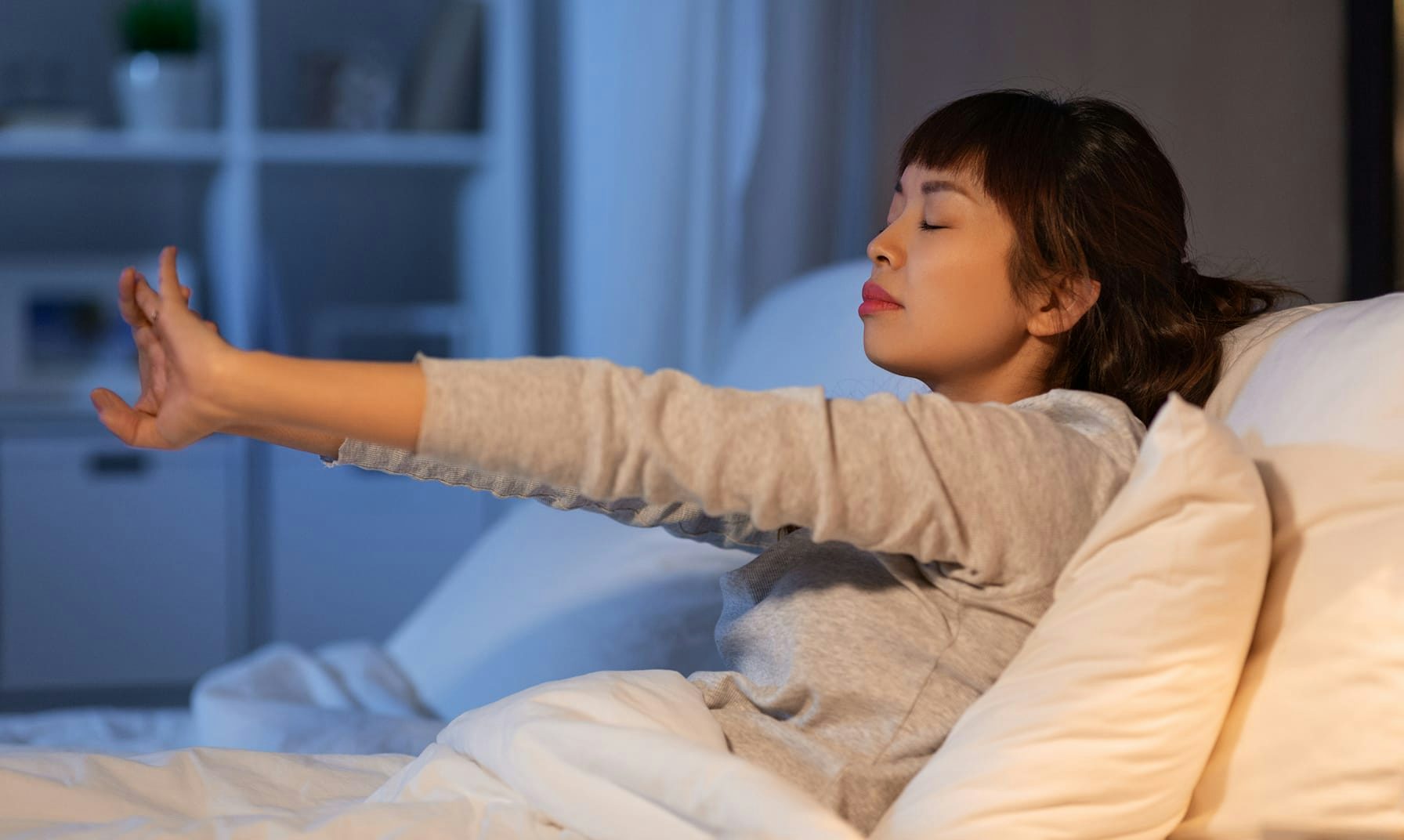 Donna asiatica che si sveglia a letto e si allunga con gli occhi chiusi, ambiente domestico notturno.