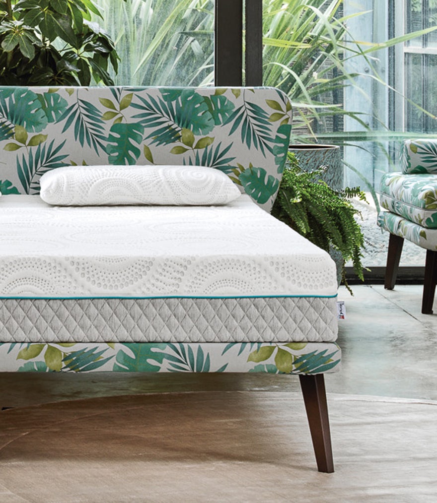 Letto con testiera e base rivestite con tessuto a foglie verdi, materasso e guanciale bianchi su pavimento chiaro davanti a…