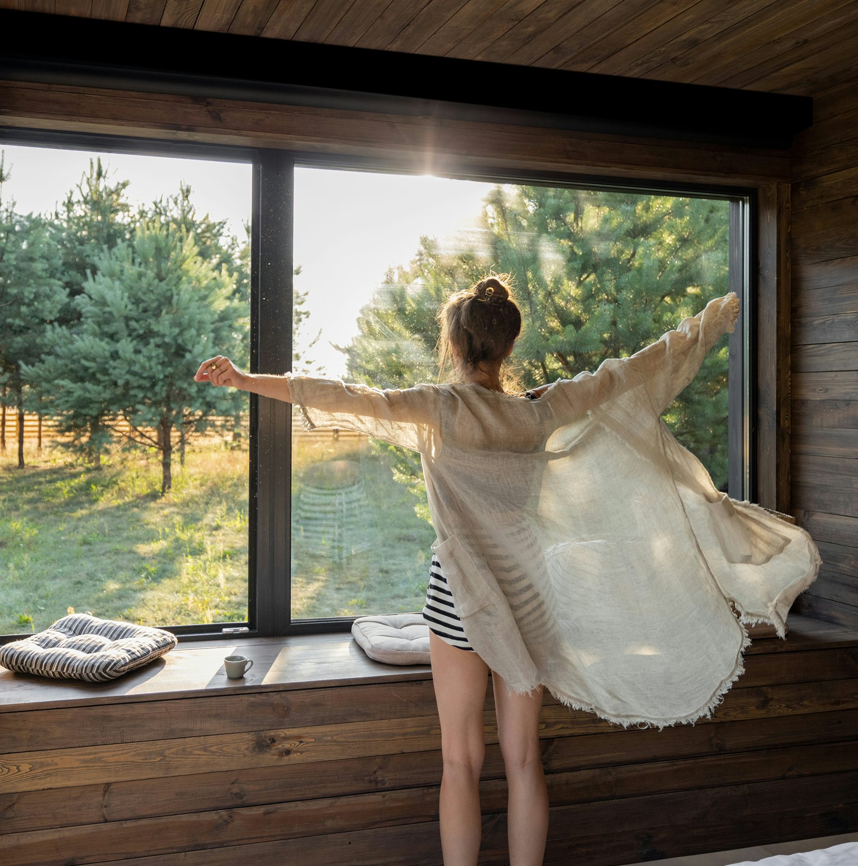 Donna con vestaglia chiara che si gode la luce del mattino davanti a una grande finestra in legno con vista su alberi verdi.