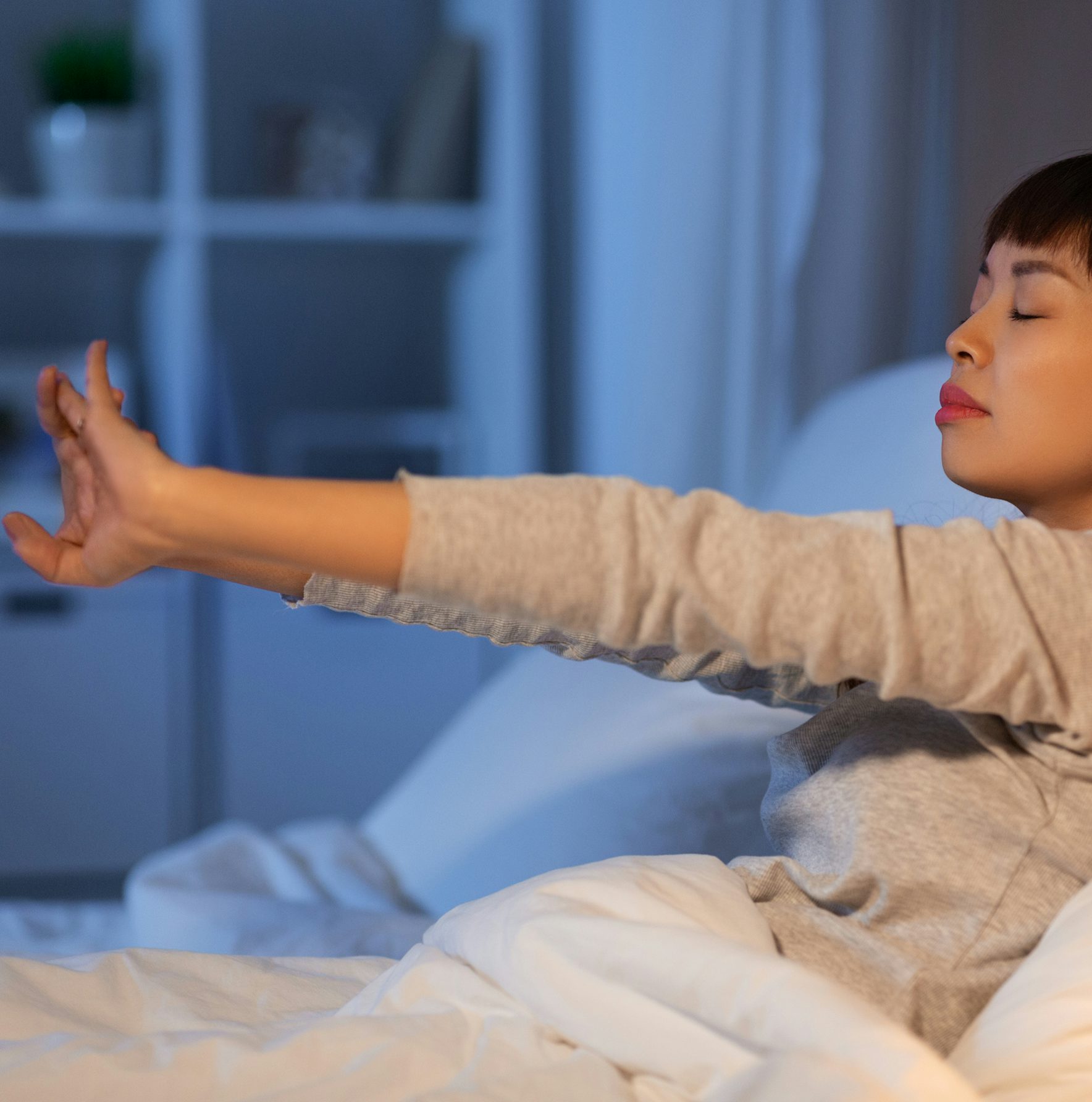 Donna asiatica con maglione grigio che si sveglia distendendo le braccia a letto con aria rilassata e occhi chiusi.