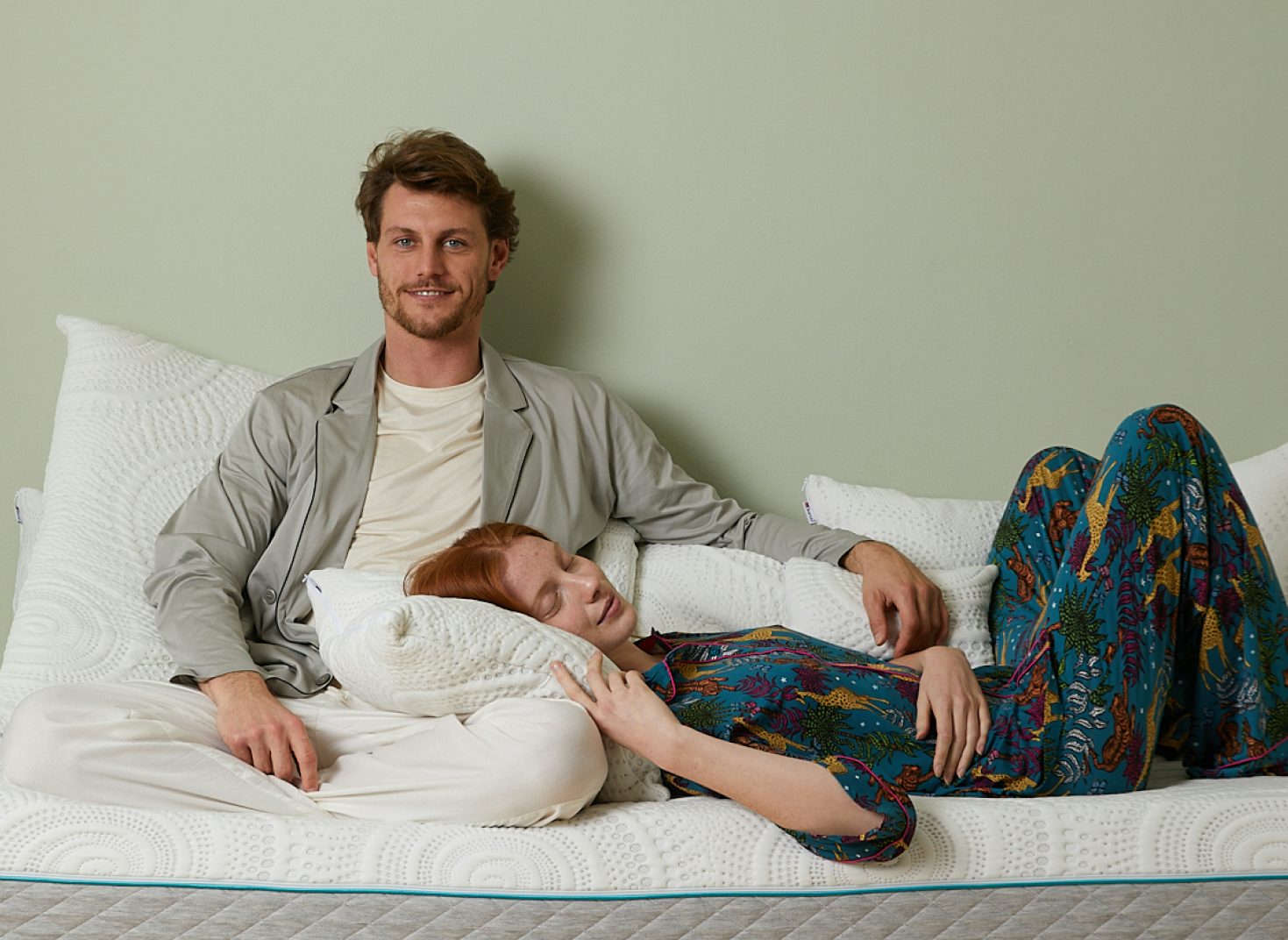 Coppia rilassata su materasso bianco, lui seduto con giacca grigia, lei con pigiama colorato sdraiata con testa sul cuscino.