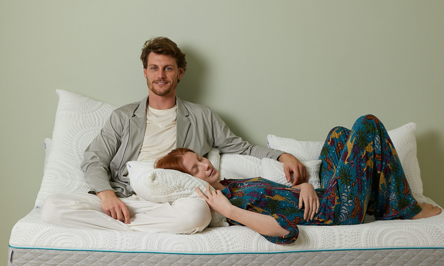 Coppia rilassata su materasso bianco, lui seduto con giacca grigia, lei con pigiama colorato sdraiata con testa sul cuscino.